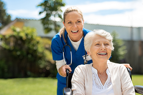 CNA in blue scrubs smiling behind elderly woman in wheelchair outside